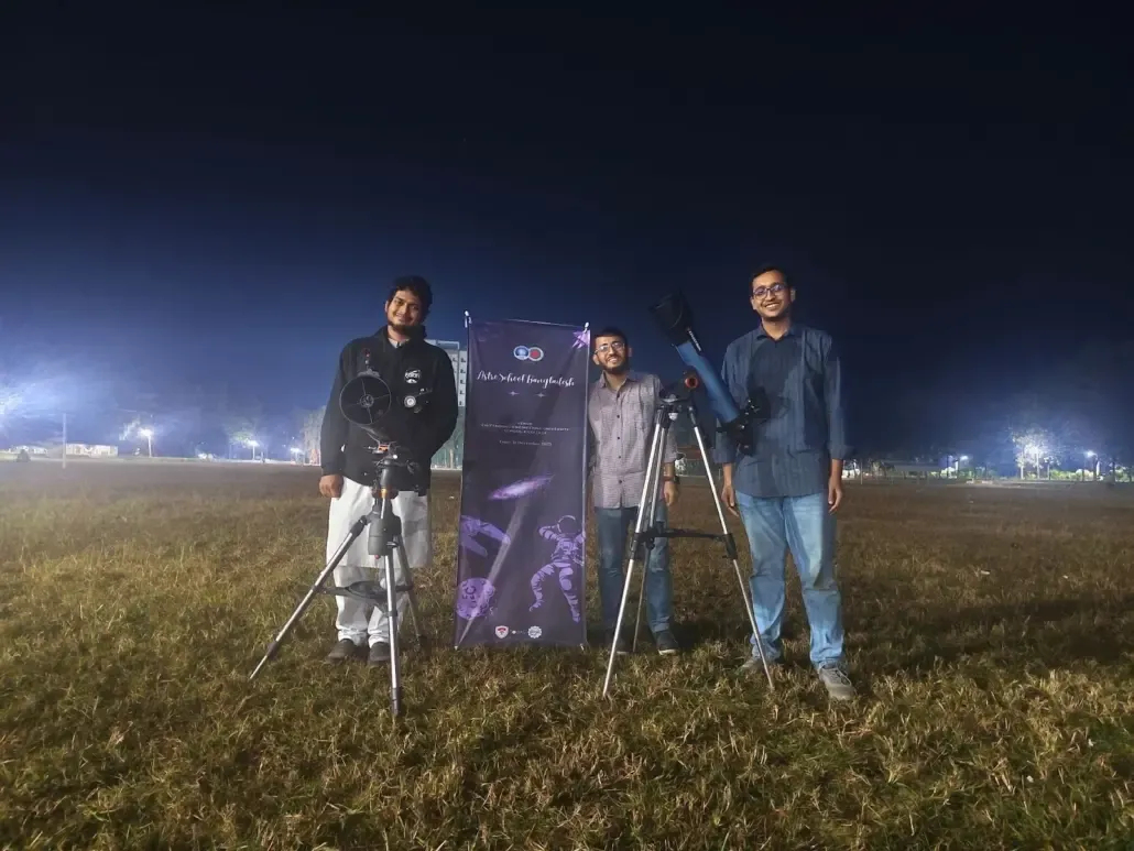 রাতের খোলা মাঠে দুটি টেলিস্কোপ ও একটি ব্যানারের পাশে দাঁড়িয়ে তিনজন স্বেচ্ছাসেবক।